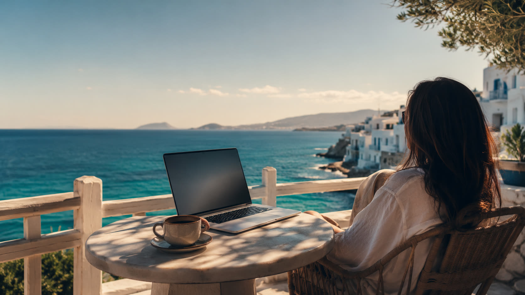 A woman works at a seaside cafe table above a turquoise Mediterranean bay.